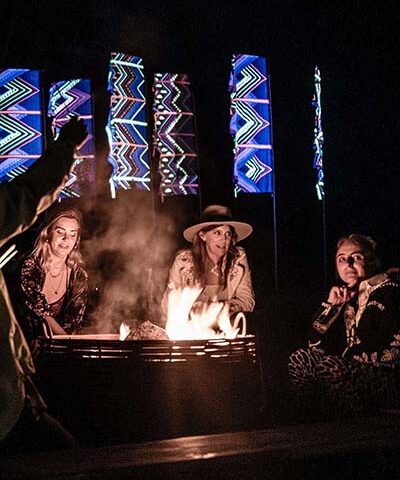 campfire scene with guests and neon flags behind